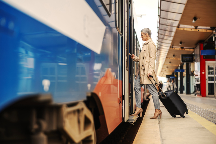 Eine ältere Frau mit einem Koffer, die in einen Zug steigt und dabei mit dem Smartphone den Fahrplan mit den Neuerungen ab Dezember 2025 checkt.